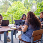 Four young women sit at wooden tables outside, working on laptops and talking. Trees and greenery surround the patio, creating a relaxed, natural atmosphere. Books and notebooks are scattered on the tables.