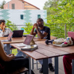 Four students sit around a wooden table outdoors, each with a laptop open. They appear to be having a discussion or study session. Green trees and modern buildings are visible in the background.