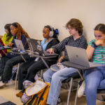 Five students sit in a classroom row working on laptops. Some wear headphones, some look focused or thoughtful. There are backpacks on the floor, and sunlight comes through a window on the left.