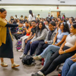 A woman in a tan cardigan interacts with seated students in a crowded room. The students are engaged, some smiling, and the atmosphere appears lively, suggesting a classroom or seminar setting.