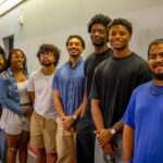 Seven young adults stand in a hallway, smiling at the camera. They are dressed casually in summer clothes, with a mix of t-shirts, shorts, and dresses. The group appears friendly and relaxed.