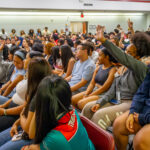 A large group of diverse students sit closely together in a classroom or auditorium. Several students have raised their hands, while others listen attentively. The room is bright and filled with engaged attendees.