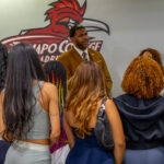 A man in a suit speaks to a group of people standing in front of him at Ramapo College, with a large bird logo and the college name visible on the wall behind him.