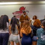 A man in a brown suit speaks to a group of students gathered around him in front of a wall with the Ramapo College Roadrunners logo. The students listen attentively, standing in a semi-circle.