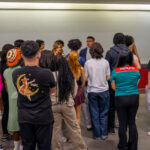 A diverse group of young adults stands in a semi-circle indoors, listening attentively to someone speaking at the front of the group. The setting appears to be a classroom or meeting space with a white wall and framed items.
