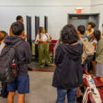 A woman speaks to a group of students gathered around her in a classroom. Most students have backpacks. There are flowers on a table, and several chairs are stacked against the wall near an exit sign.