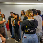 A group of young adults stands in a semi-circle, attentively listening to a man speaking. The setting appears to be indoors, possibly at a casual event or workshop. Some are holding drinks and flowers are seen on a table.