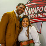 A man in a mustard suit and a woman in a white blouse and green skirt stand smiling in front of a Ramapo College Roadrunners sign on a wall.