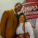 A man in a brown suit and a woman in a white blouse with a brown belt smile together in front of a Ramapo College Roadrunners sign.