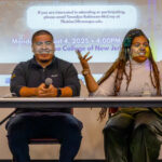 Two people sit at a table speaking in front of a projected screen with event information. The person on the left is smiling with hands folded, while the person on the right gestures animatedly with both hands raised.
