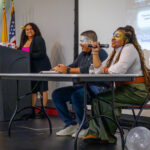 Three people sit and speak at a table on a stage while another stands at a podium. A projector screen and U.S. flags are visible in the background. The atmosphere appears to be that of a panel discussion or presentation.