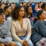 A diverse group of students sits closely together in an auditorium, attentively listening to a speaker. Some wear glasses, and their expressions range from focused to curious.