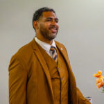 A man in a brown suit and striped tie stands smiling beside a vase of orange flowers, with a light gray background.