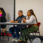 Three people are in a room: one woman stands at a podium while a man and another woman sit at a table with microphones in front of them, appearing to participate in a discussion or panel event. An American flag is in the background.