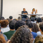 A woman speaks passionately at a table beside a man during a panel discussion, while an audience faces them, and another woman stands at a podium to the side.