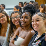 Four young women sit closely together, smiling at the camera in a crowded indoor setting, with other people blurred in the background.