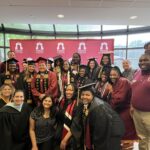 A group of graduates in caps and gowns pose and smile with faculty members in front of a pink banner that reads Ramapo College inside a bright, windowed room.