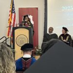 A student in graduation attire speaks at a podium on stage, with the American flag, officials in academic regalia, and a large screen displaying “2025” and “Ramapo College” in the background. Audience members are visible in the foreground.