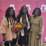 Three women stand smiling in front of a Ramapo College of New Jersey backdrop. The center woman wears a graduation cap and gown, holding flowers and an award, while the two women beside her wear patterned dresses.