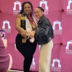 Two women smile and point toward the camera in front of a pink Ramapo College of New Jersey backdrop. One wears a graduation stole, medals, and cords; the other wears light pants and a dark jacket.