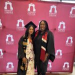 Two women stand smiling in front of a Ramapo College of New Jersey backdrop. One wears a graduation cap, gown, and multiple stoles, while the other wears academic regalia with a red and black sash.