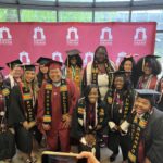 A group of smiling graduates in caps and gowns pose together indoors in front of a Ramapo College banner. Most wear colorful stoles, and the mood is joyful and celebratory.
