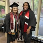 Two women wearing graduation gowns and caps stand smiling indoors in front of a Ramapo College backdrop. One graduate is wearing multiple honor cords, stoles, and medals while the other is in a black gown with a red and white stole.
