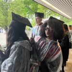 Graduates in caps and gowns stand under a shelter on a rainy day, wearing clear plastic ponchos. One person in the foreground smiles and makes a peace sign while others face away or look forward. Green trees are visible outside.