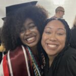 Two women smiling and hugging at a graduation ceremony. One is wearing a graduation cap, gown, honor cords, and a sash; the other is in a black robe. They appear happy and excited, with people in the background.