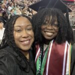 Two women smiling at a graduation ceremony, one wearing a graduation cap, gown, and honor cords, the other also in a black gown, with a crowd of people seated in the background.