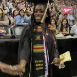 A smiling graduate in cap and gown, wearing a colorful kente stole and a sash with her name Esther Adwoa Oduro, stands in front of a crowd at a graduation ceremony. She holds a yellow paper in one hand.