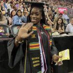 A smiling graduate in a cap and gown waves at the camera. She wears a kente stole with the name “Esther Adwoa Oduro.” The background shows a seated, cheering audience and a “School of Business” sign.