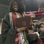 A smiling graduate wearing a cap and gown holds a diploma folder at a commencement ceremony in an indoor arena, with other graduates seated nearby and an audience in the background.