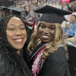 Two women in graduation gowns and caps smile for a selfie at a commencement ceremony, surrounded by an audience seated in an arena. One wears honor cords and a lei.
