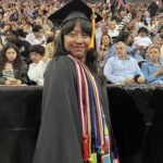 A smiling graduate in a cap and gown stands proudly with colorful honor cords, in front of an audience at a graduation ceremony. The sign above reads School of Contemporary Arts.