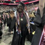 A smiling graduate in a cap and gown waves at the camera, wearing multiple honor cords and stoles. She stands among other graduates at a large indoor commencement ceremony, with a crowd seated in the background.