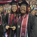 Two graduates in caps and gowns smile at the camera during a commencement ceremony at Ramapo College of New Jersey. They wear maroon stoles and medals, with a crowd of spectators seated behind them.