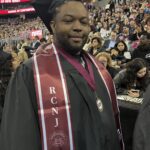 A graduate wearing a black cap and gown and a maroon stole labeled RCNJ stands in an arena filled with seated spectators. The person also wears a graduation medal around their neck.