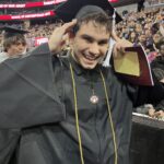 A young graduate in a black cap and gown smiles widely while holding his graduation cap. He wears a gold honor cord and a medallion. The background shows a crowd and digital signs for Ramapo College’s graduation ceremony.