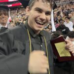 A young graduate in a cap and gown smiles joyfully at a graduation ceremony, holding a diploma folder. A crowd of people and other graduates are visible in the background.