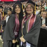 Two graduates in caps and gowns smile for a photo at a commencement ceremony. They wear maroon stoles and medals, standing in front of a large seated audience and digital signs reading Ramapo College of New Jersey.