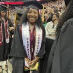 A smiling graduate in a cap and gown stands among a crowd at a commencement ceremony. She wears a white stole with First Generation Graduate written on it, and the arena is filled with people in the background.