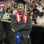 A graduate in a cap and gown, wearing a stole with the U.S. flag and a blue stole with a white star, smiles with arms crossed at a graduation ceremony. The audience is seated in the background.
