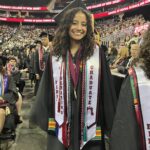 A smiling graduate wearing a cap and gown stands in an arena. Her sash reads First Generation Graduate. Rows of graduates and audience members are seated behind her. Red banners display Ramapo College of New Jersey.