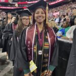 A smiling graduate in cap and gown stands indoors at a ceremony with others in similar attire. Her sash reads “RCNJ” and “EOF Black, Civil, Magic.” Audience members are seated in the background.
