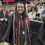 A smiling graduate in a cap and gown stands at a commencement ceremony. She wears honor cords, a medal, and a stole that says EOF. A crowd and Ramapo College of New Jersey banners are visible in the background.