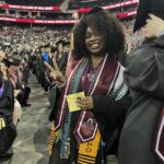 A smiling graduate wearing a cap and gown stands among other graduates, holding a piece of paper and making a peace sign. She is adorned with several honor cords and stoles. The venue is filled with people.