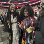 Two graduates wearing caps and gowns smile at the camera during a graduation ceremony. One holds up a peace sign and both wear multiple honor cords and stoles, with a crowd and banners visible in the background.
