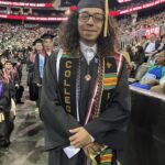 A graduate in a cap and gown stands smiling at a commencement ceremony in an arena, with a crowd and other graduates in the background. He wears multiple cords, sashes, and a stole embroidered with COLLEGE.
