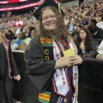 A smiling graduate in a cap and gown stands in front of a crowd at a graduation ceremony. The student wears several honor cords and sashes, including one that says First Generation. Ramapo College banners are visible in the background.